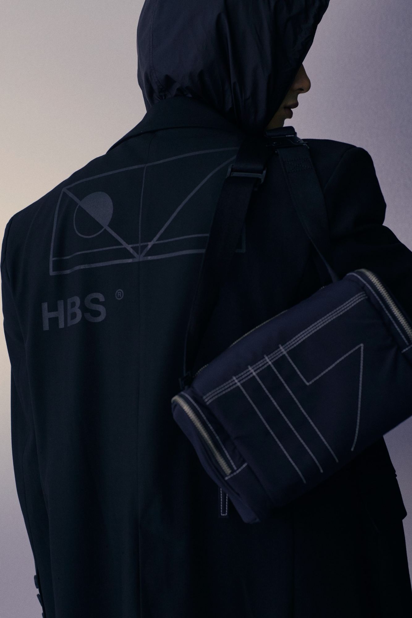 Lưng áo khoác đen in hình học và chữ HBS, đeo túi đen
